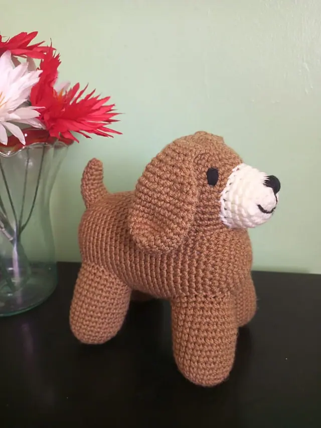 Brown crocheted dog with floppy ears, a cream muzzle and black nose, standing on a dark table in front of a pale green wall, with red and white flowers in a glass vase at the left.