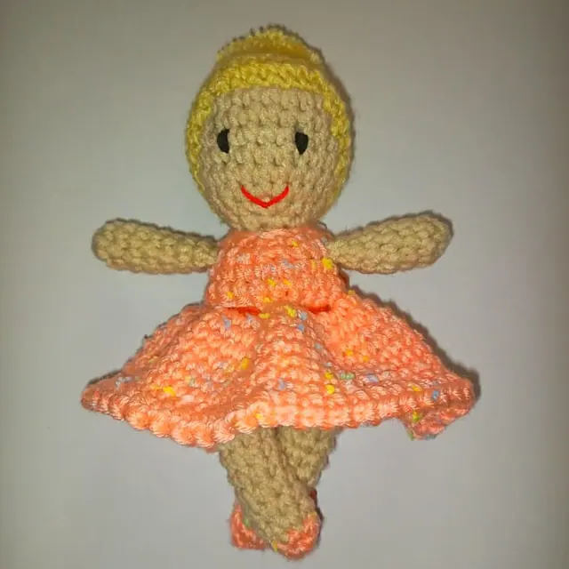 Small crocheted doll with yellow yarn hair, beige face, black eyes and a red stitched smile, wearing a peach flecked crocheted dress.