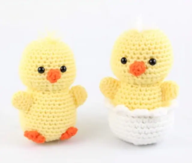 Two yellow crocheted ducklings side by side with orange beaks and feet, one standing and one sitting in a white crocheted eggshell.