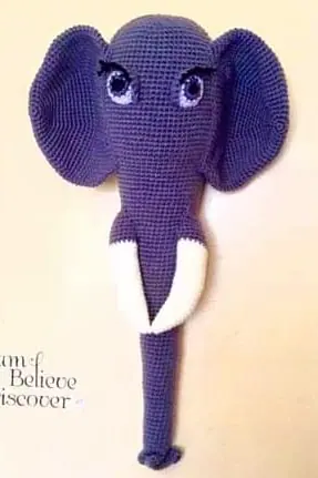 Crocheted purple elephant head and long trunk on a cream wall, with large ears, lilac eyes with lashes and white tusks.