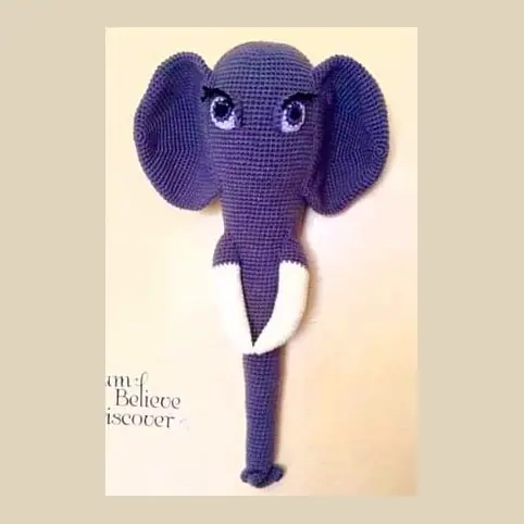 Crocheted purple elephant head and long trunk on a cream wall, with large ears, lilac eyes with lashes and white tusks.