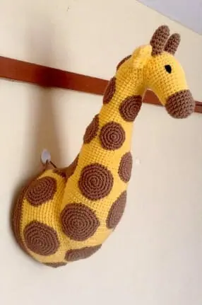 Crocheted yellow giraffe neck and head mounted on a wall rail, covered in round brown spots with two small horns and a brown muzzle.