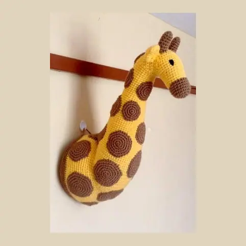 Crocheted yellow giraffe neck and head mounted on a wall rail, covered in round brown spots with two small horns and a brown muzzle.