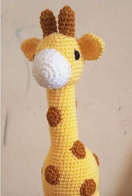Crocheted yellow giraffe with brown spots, white muzzle and brown horns, head and neck against a plain cream wall