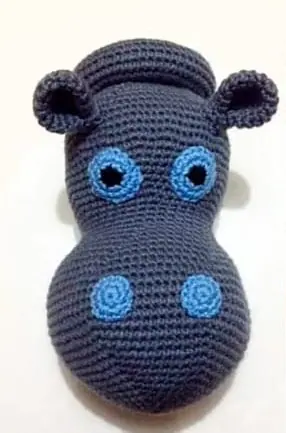 Crocheted dark grey hippo head facing front on a cream wall, with small round ears, blue ringed eyes and two blue nostril circles.