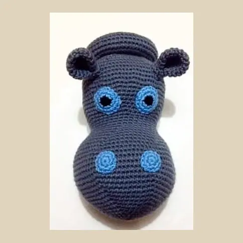 Crocheted dark grey hippo head facing front on a cream wall, with small round ears, blue ringed eyes and two blue nostril circles.