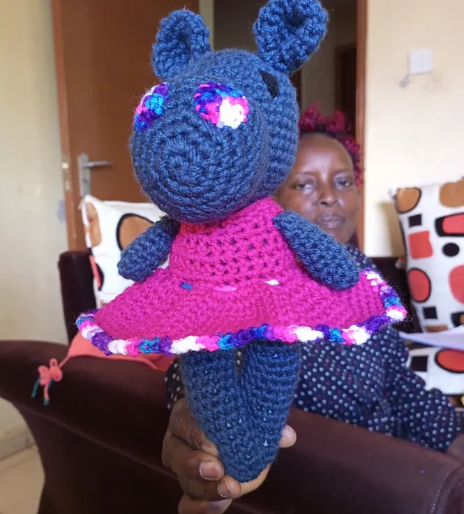 Navy crocheted hippo in a magenta crocheted dress with a multicoloured hem, held up in a hand, with a woman seated on a sofa behind
