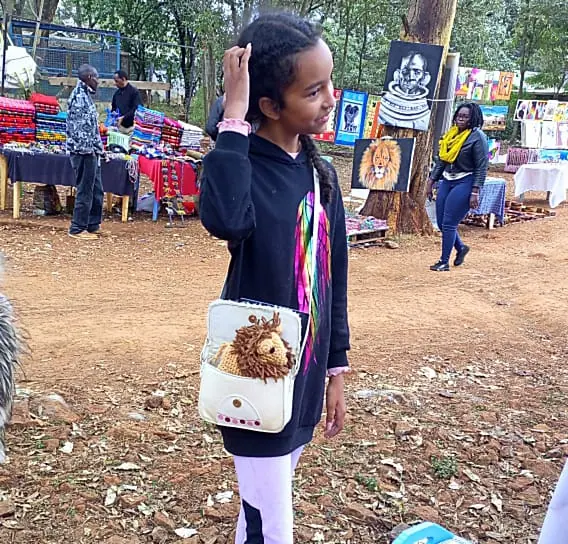 A white shoulder bag worn across the body, with a crocheted lion head with a brown yarn mane looking out of the opening