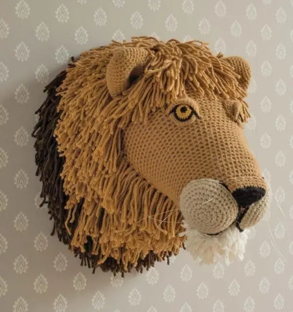 Crocheted tan lion head mounted on a patterned grey wall, with a long fringed golden mane edged in dark brown, yellow eyes, cream muzzle and black nose.