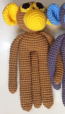 Long limbed brown crocheted monkey with a yellow face and ears, black eyes and a yellow muzzle, with the arm of the next monkey at the edge