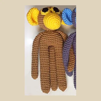 Long limbed brown crocheted monkey with a yellow face and ears, black eyes and a yellow muzzle, with the arm of the next monkey at the edge