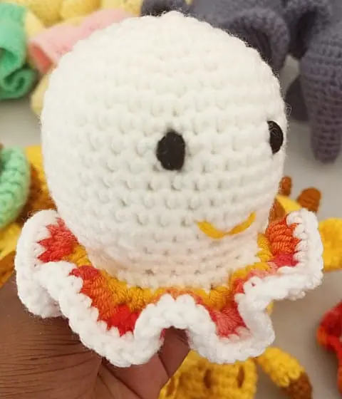 Hand holding a white crocheted octopus with a ruffled skirt edged in white and shaded red, orange and yellow, with black eyes and a yellow smile