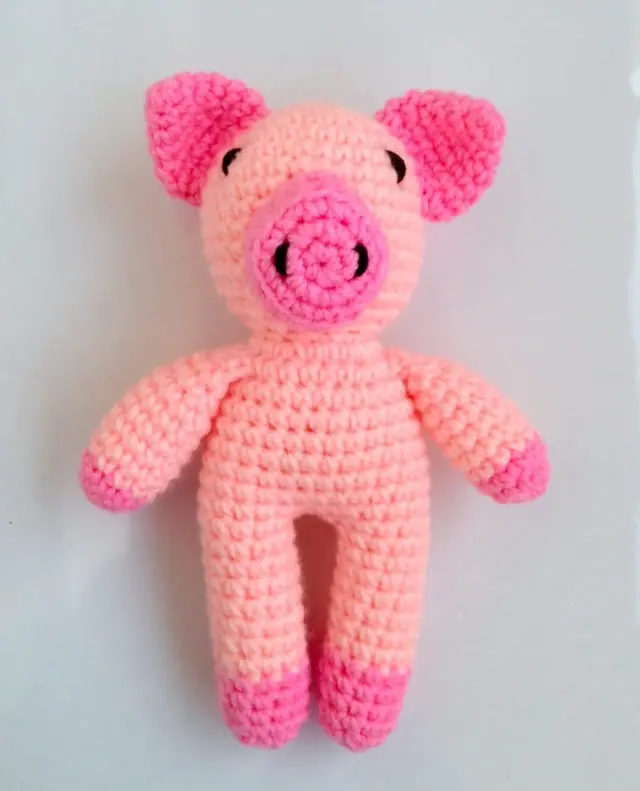 Pink crocheted pig standing upright, with a peach body, bright pink ears, snout and hooves and small black eyes, on a pale grey background.