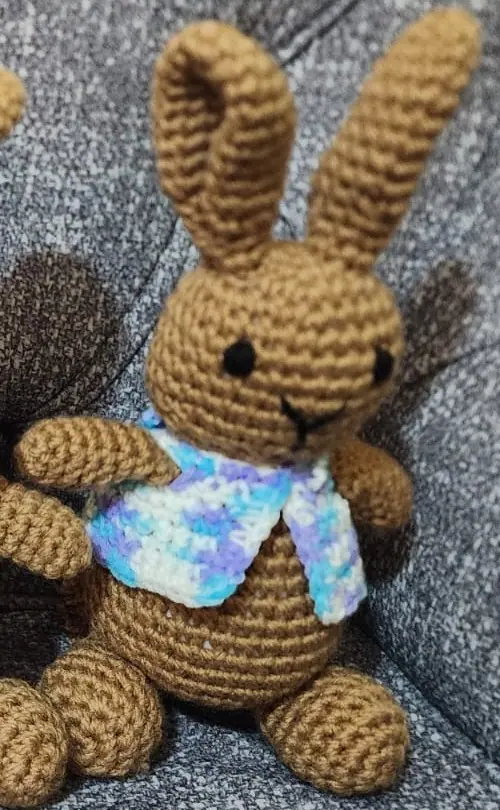 Brown crocheted rabbit with one folded ear wearing a vest in pale blue, lilac and white, sitting on a grey sofa.