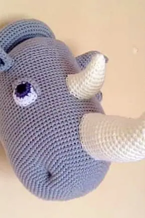 Crocheted grey rhino head seen from the side on a cream wall, with a lilac eye, a cream front horn and a smaller cream horn.
