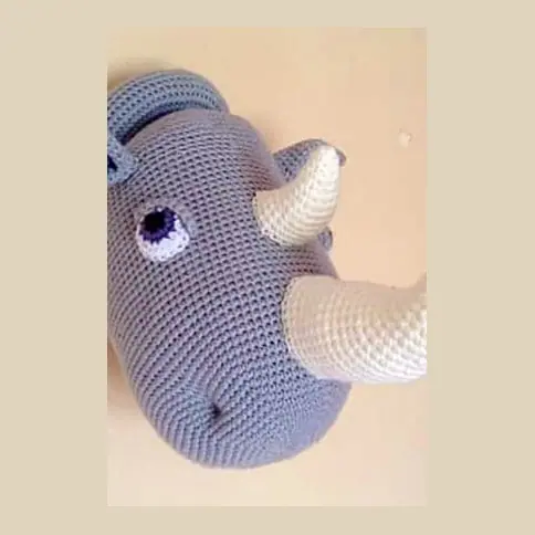 Crocheted grey rhino head seen from the side on a cream wall, with a lilac eye, a cream front horn and a smaller cream horn.