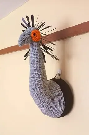Crocheted grey secretary bird neck and head mounted on a round black plaque on a wall rail, with an orange face patch and black crest feathers.