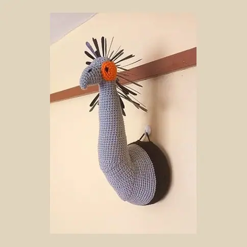 Crocheted grey secretary bird neck and head mounted on a round black plaque on a wall rail, with an orange face patch and black crest feathers.