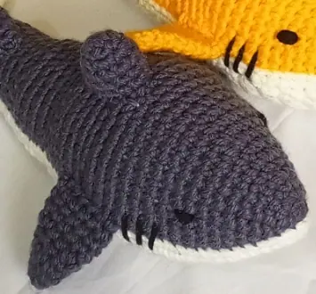 Dark grey crocheted shark with a cream underside, a black eye and stitched gill lines on a white sheet, with the edge of the next shark in the frame