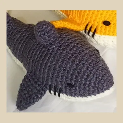 Dark grey crocheted shark with a cream underside, a black eye and stitched gill lines on a white sheet, with the edge of the next shark in the frame