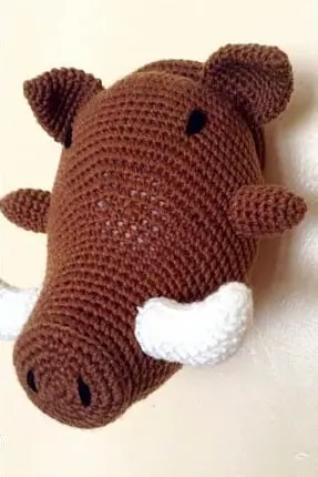 Crocheted brown warthog head on a cream wall with small ears, two white curved tusks and dark eyes and nostrils.