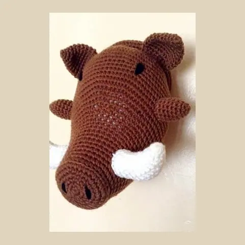 Crocheted brown warthog head on a cream wall with small ears, two white curved tusks and dark eyes and nostrils.