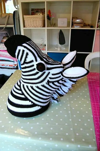 Crocheted black and white striped zebra head standing on a spotted tablecloth with a shelving unit behind, showing a spiky mane, round dark eye and black muzzle.