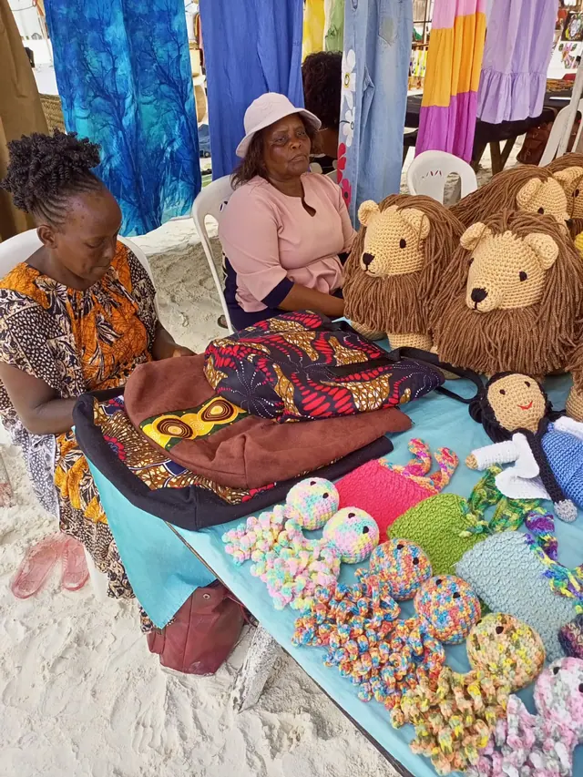 Two women at a stall on sand with crocheted lions and dolls on a blue table
