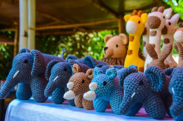 Blue and grey crocheted elephants and rhinos in front of giraffes and a brown bear under a bamboo roof