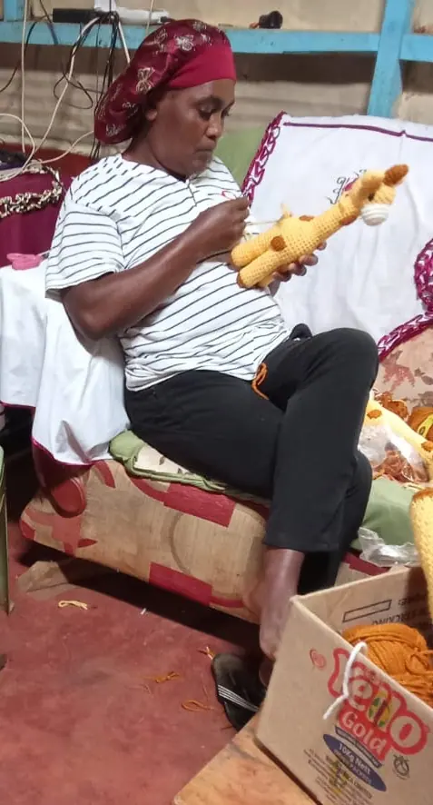 Woman in a striped top and headscarf holding a crocheted yellow giraffe, with a box of orange yarn beside her