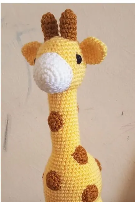 Crocheted yellow giraffe with brown spots, a white muzzle and two brown horns against a cream wall