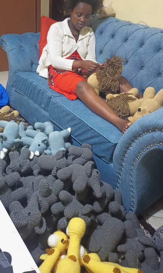 Woman in a white jacket on a blue sofa working on a lion beside a pile of grey crocheted elephant pieces