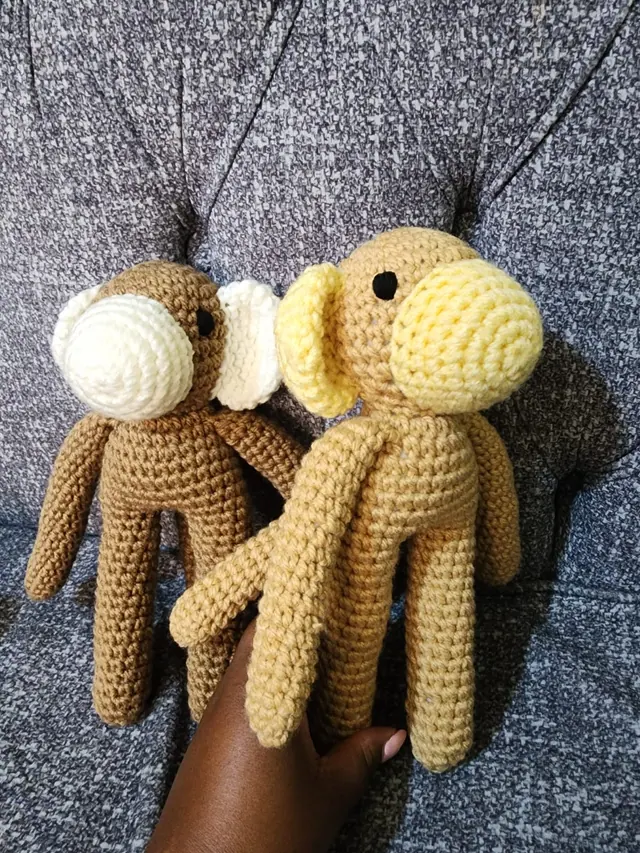 Two long-limbed crocheted monkeys, one brown with cream ears and one tan with yellow ears, held by a hand against a grey sofa