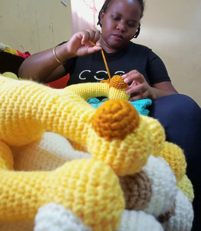 Woman in a black t-shirt crocheting with a hook, with yellow and cream crocheted giraffe pieces in the foreground