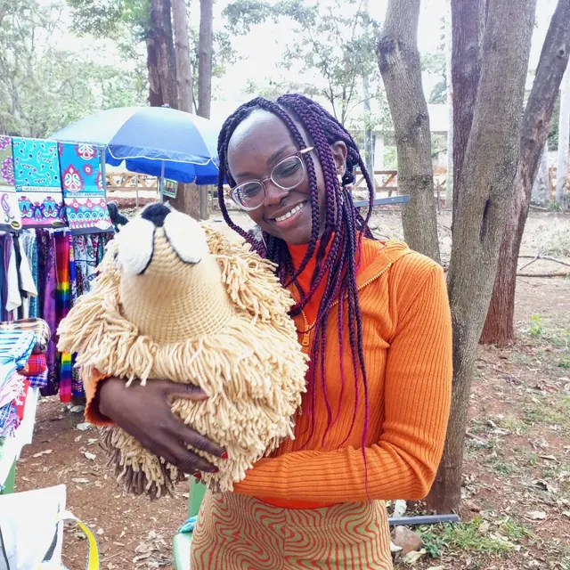 Smiling woman with purple braids and an orange top holding a large crocheted lion at an outdoor market with kitenge cloths behind her