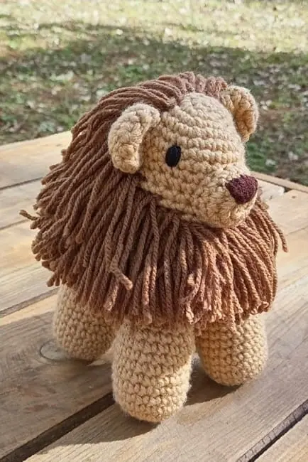 Crocheted lion with a tan body and a long brown yarn mane standing on a wooden pallet in the sun