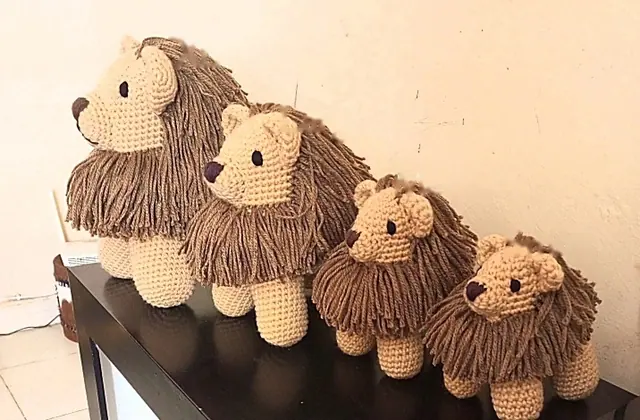 Four crocheted tan lions with brown manes in a row on a black table, from largest at the left to smallest at the right, against a cream wall