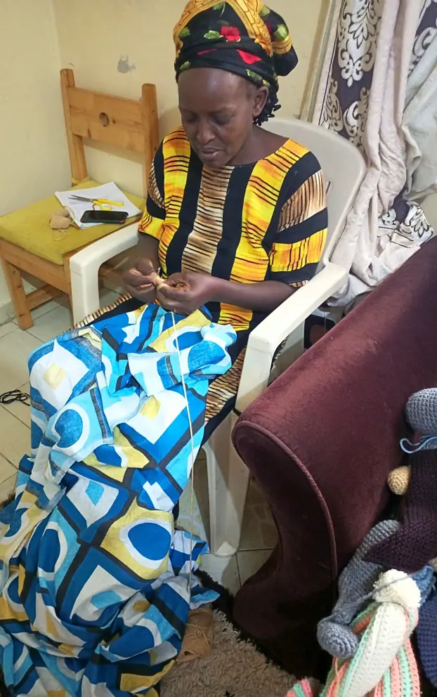 Woman in a patterned yellow and black top and kitenge skirt crocheting on a chair, with a crocheted animal waiting beside her