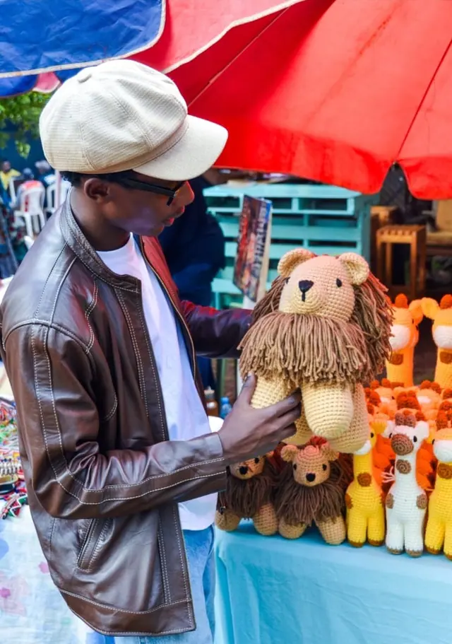 Man in a cap and leather jacket looking closely at a crocheted lion at a market stall