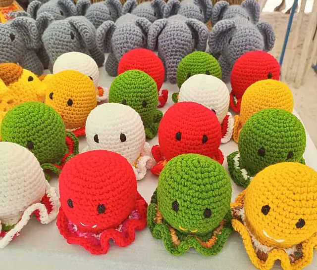 Rows of red, green, white and yellow crocheted octopuses in front of grey crocheted elephants on a white table.