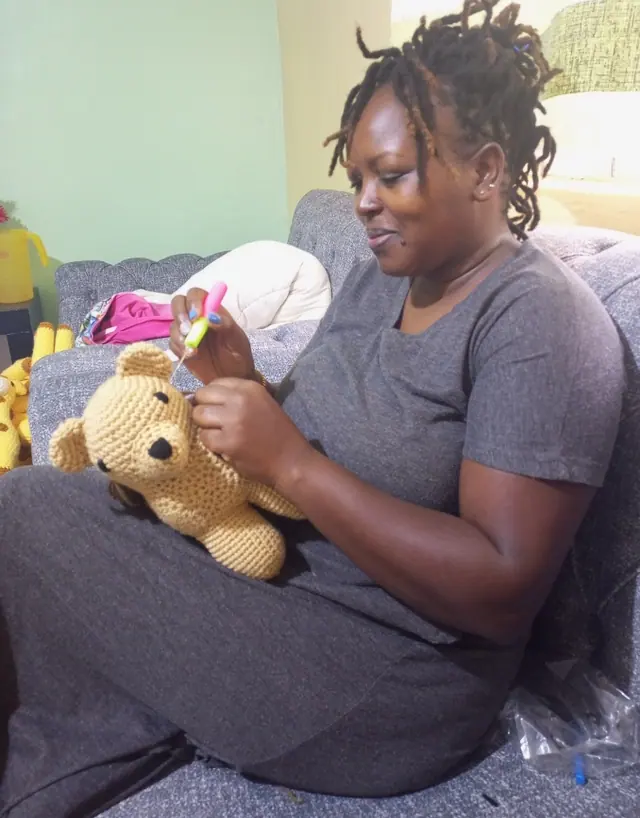 Woman with locs on a grey sofa sewing the face of a tan crocheted bear on her lap