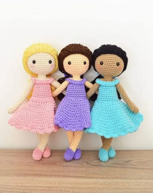 Three crocheted dolls in pink, lilac and turquoise dresses standing against a white wall