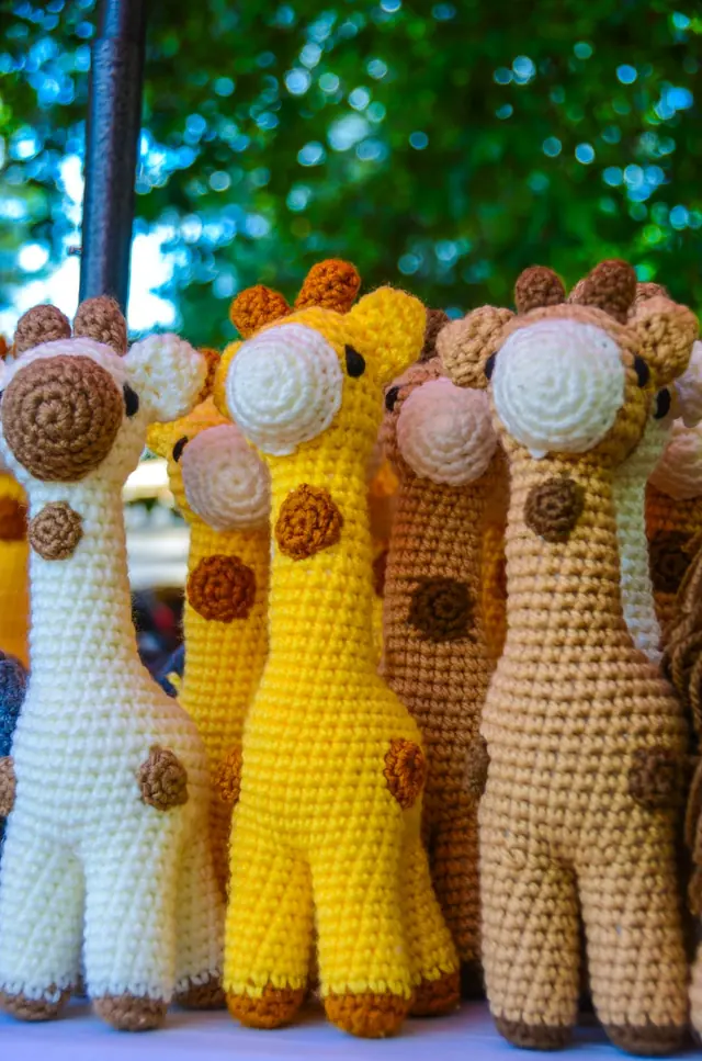 Row of crocheted giraffes in cream, yellow, brown and tan with spiral spots, standing on a stall under trees