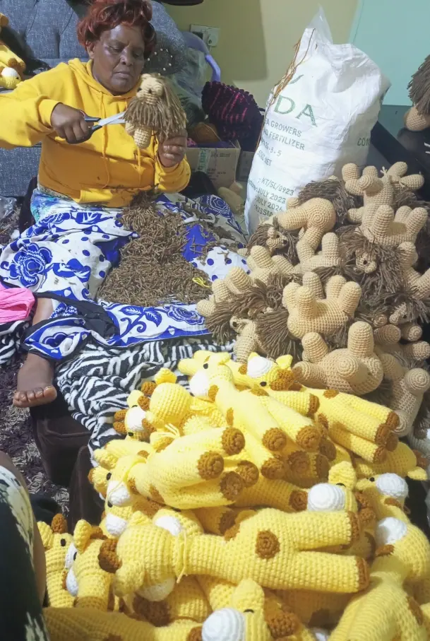 Woman in a yellow hoodie trimming a lion mane with scissors among piles of crocheted giraffe and lion pieces